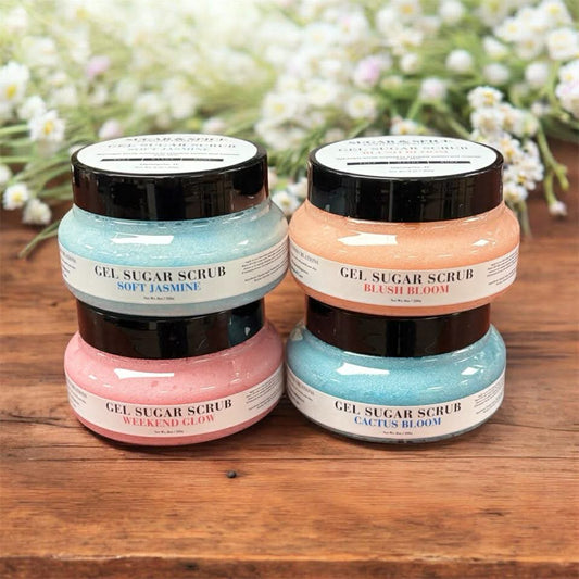 Four jars of gel sugar scrub with different scents on a wooden surface with flowers in the background.